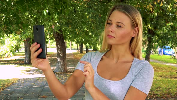 Young Pretty Blond Woman Takes Photos Wirh Smartphone - Park with Trees in Background