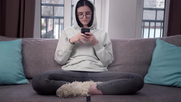 Pretty Girl in a Hood and Glasses Uses a Cell Phone Sitting at Home on the Couch Window Background