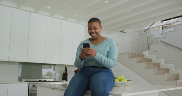 Happy african american plus size woman sitting on countertop in kitchen, using smartphone