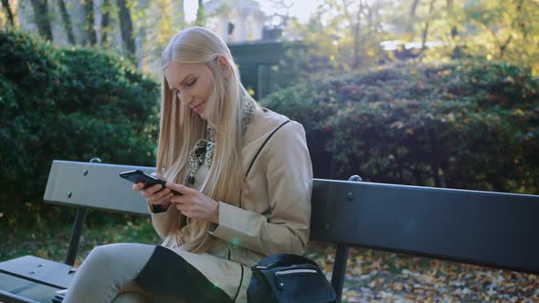 Young Blonde Girl Uses a Cell Phone on a Bench in the Park Outdoors.