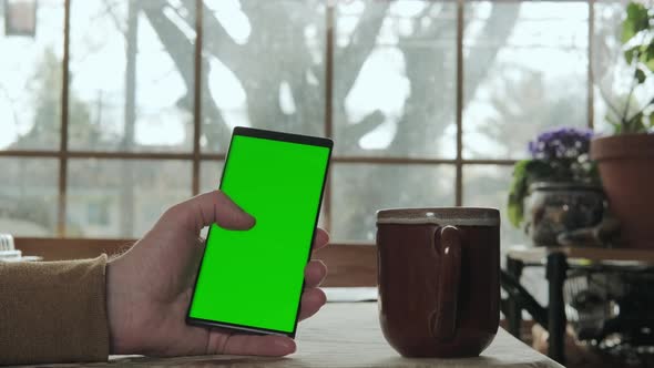 A Male Hand Holds a Phone with a Mockup Green Screen and Drinks Coffee