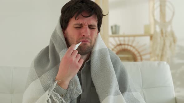 Man with a Cold is Sitting on the Couch with a Thermometer in His Mouth