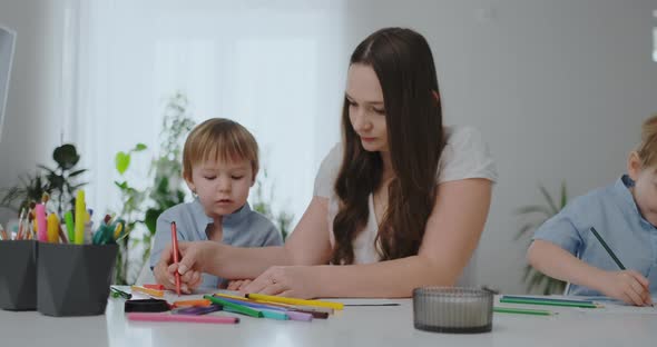 A Young Mother with Two Children Sitting at a White Table Draws Colored Pencils on Paper