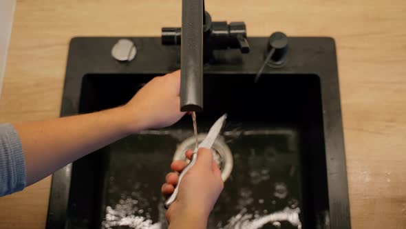 Unrecognizable person washing steel knife in sink closeup. Wooden table and mans hands are visible