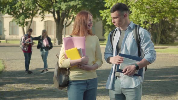 Portrait of Positive Caucasian Boy and Girl Talking on University Campus Yard with Blurred Students