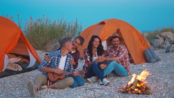 Picnic of Young Adult People with Bonfire and Ukulele on Beach with Tents at Evening