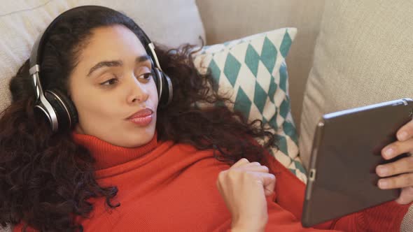 Happy woman lying on sofa using tablet enjoying music on her headphones 4K 4k