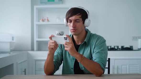 Young Man in Headphones Playing App Game on His Smartphone While Sitting on Kitchen Table at Home