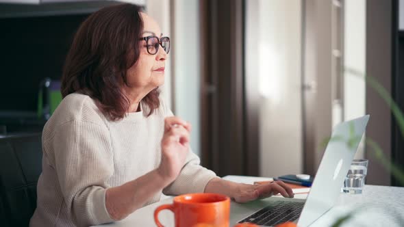A Senior Woman in Glasses Working at Home with a Laptop and Drinking Coffee