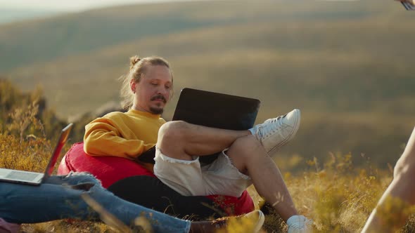 Caucasian Man Hoodie and Shorts is Sitting an Easy Chair in Nature with a Laptop on His Lap Front