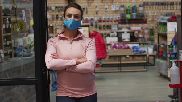 Caucasian female shopkeeper wearing face mask leaning in the doorway of sports shop