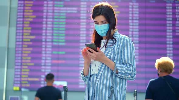 Female Air Passenger in Mask, Standing Against Departure Board at Airport. She Is Checking Flight