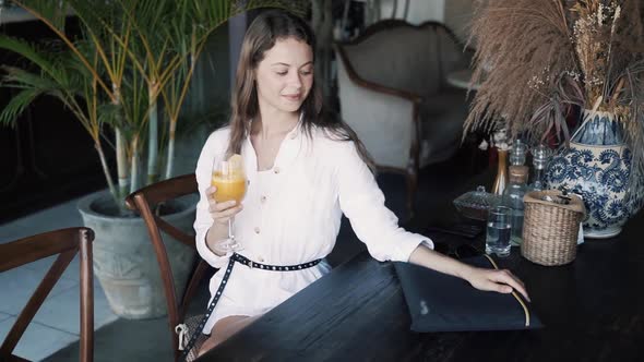 Girl Sits at Table in Cafe and Holds in Her Hands Glass with Orange Juice