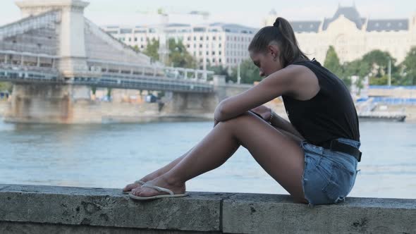 Young Woman Uses Smartphone Outdoor on the Embankment in the Park Near River