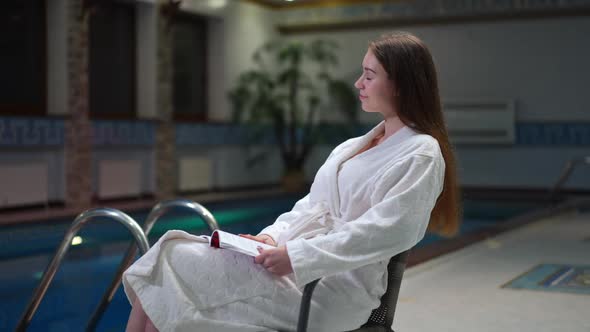 Side View Young Happy Woman Dreaming Sitting with Book at Pool Indoors Smiling