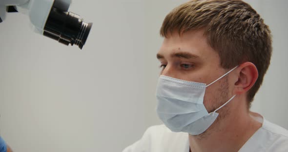 A Male Doctor or Scientist in a Medical Mask Looks Through a Microscope