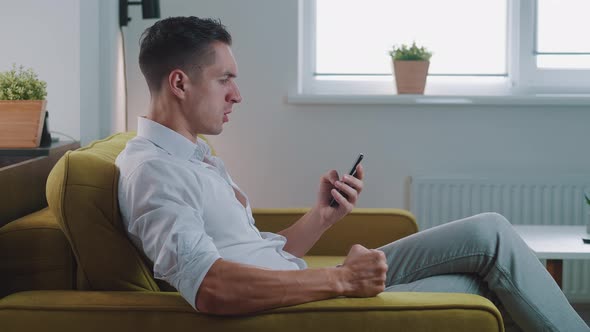 Annoyed Young Man Holding Smartphone Frustrated By Bad Message, Sitting on Sofa at Home. Angry Male
