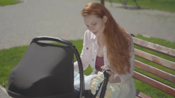 Portrait Red-haired Woman Playing with Her Child Sitting on the Bench. The Stroller Standing Near