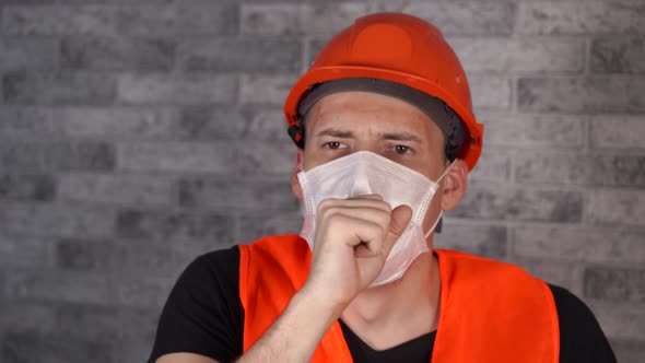 Male Construction Worker in Overalls and Medical Mask Coughing on Face Background of Gray Brick Wall