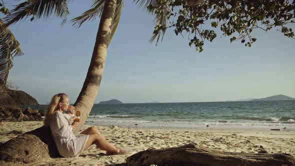 Woman Sitting in White Dress Drinks Pineapple Cocktail Pina Colada on Beach Near Palm