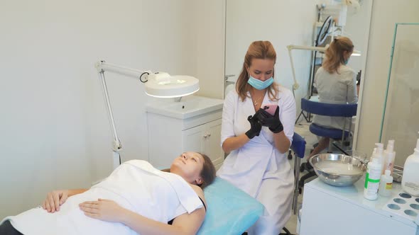 Cosmetologist in White Coat is Sitting in Office and Typing Text on Smartphone