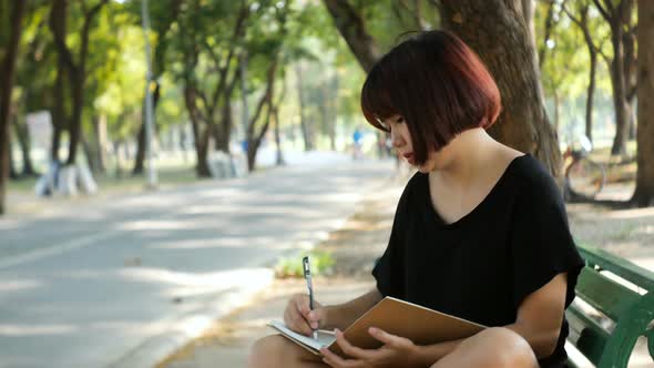 young asian woman writing into her diary working on notebook in park. Student studying outdoors.