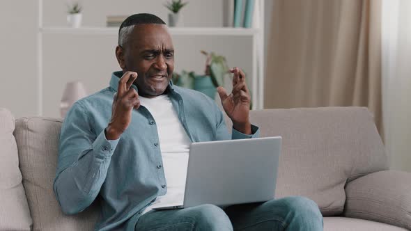 Mature African American Man Sitting in Room Fingers Crossed Hopefully Looking at Laptop Screen Asks