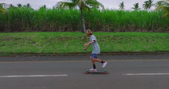 A Teenager Rides a Longboard Along a Beautiful Road with Green Palm Trees