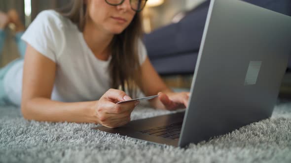 Woman with Glasses Is Lying on the Floor and Makes an Online Purchase Using a Credit Card and Laptop