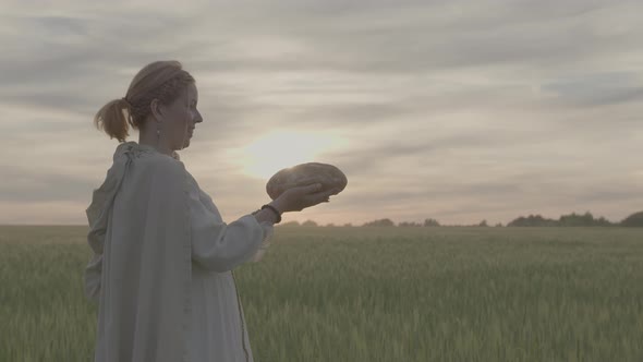 Young Woman with a Loaf of Fresh Bread in Her Hands Stands in a Field at Sunset
