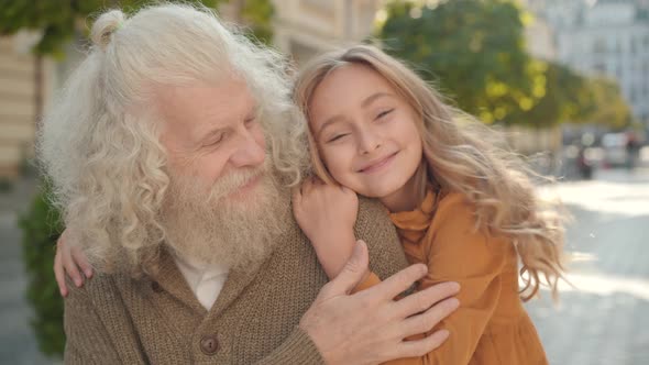 Close-up Portrait of Happy Grandfather and Granddaughter Posing in Sunlight Outdoors. Cheerful