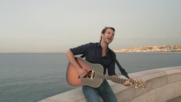 A Good Looking Man Joyfully Singing and Playing His Guitar with a Beautiful Bay in the Background, M