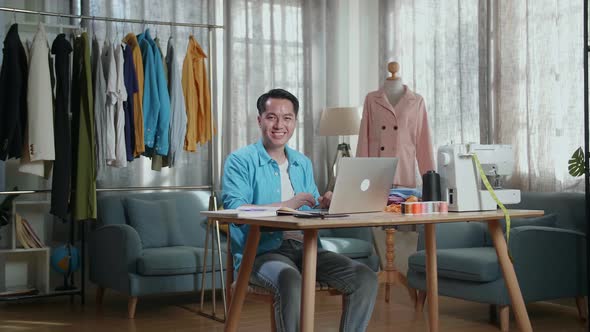 Asian Male Designer Working On A Laptop And Smiling To The Camera While Designing Clothes