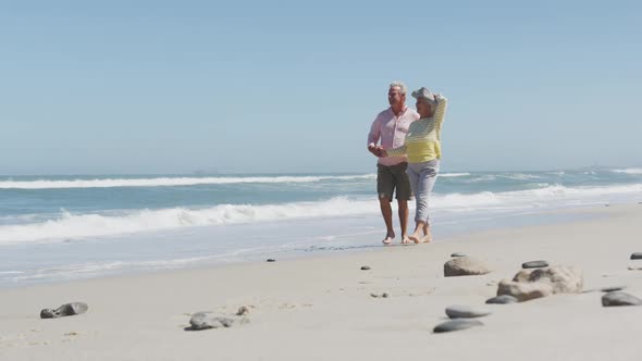 Happy senior caucasian couple holding hands talking to each other while walking on the beach