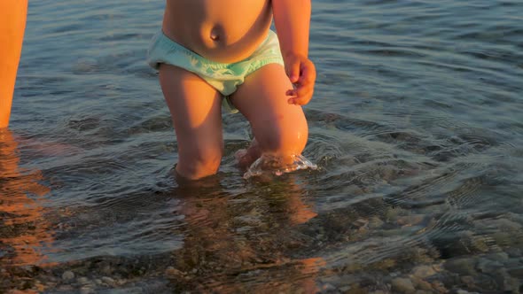 Mom Helps To Make the Child's First Steps on the Beach. Baby Doing First Steps in Sea Water