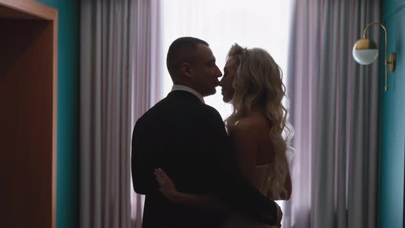 Young Man Kisses Nose of Blonde Curly Haired Woman By Window