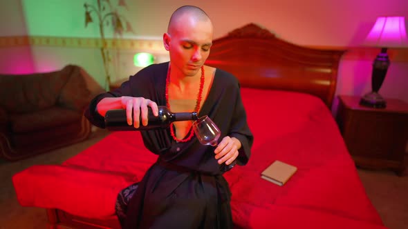 Young Man Pouring Wine in Glass Sitting on Bed in Red Lights