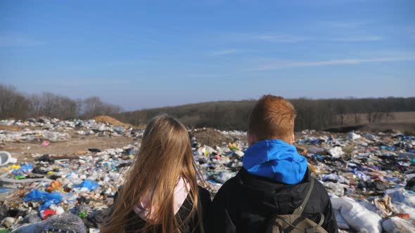 Rear View of Young Girl and Boy Stands Against the Blurred Background of Dirty Garbage Dump