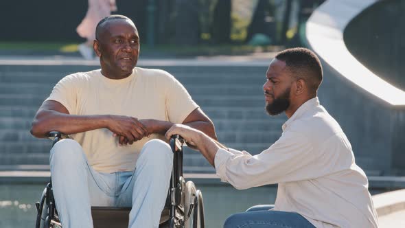 Young African American Guy Squatting Down Talking to Elderly Father in Wheelchair