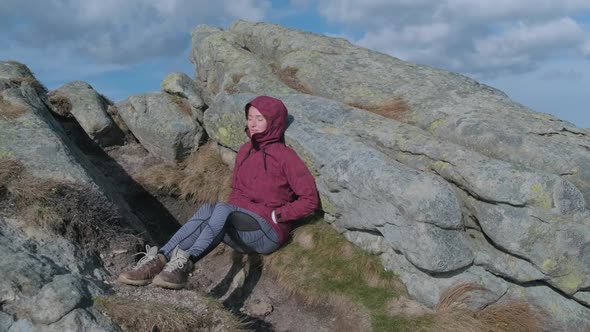 Exhausted Tourist Woman After Hard Trekking in Mountains Sits Near Large Rock Relaxes in Sun