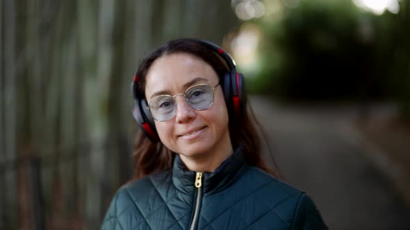 Portrait of a Smiling Adult Woman Wearing Glasses and Headphones Listening to Music in a City Park