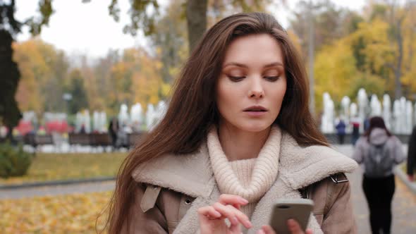 Pivoting Portrait of Discompose Brunette American Woman Holding Smartphone in Autumn Park Centre and