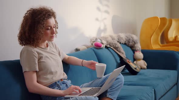 Smiling Young Woman Sitting on Couch Using Laptop Notebook Looking at Screen Typing Message Happy