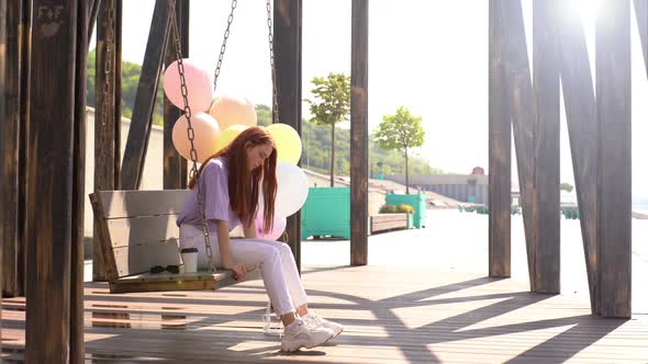 Sad Redhead Young Woman Swaying on Swing Holding Hands Large Fountain of Colorful Balloons.