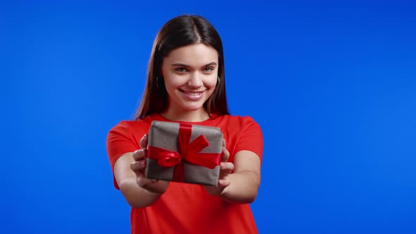 Woman Holding Gift Box and Gives It By Hands to Camera on Blue Wall Background