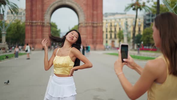 Woman Photographing Her Gorgeous Best Female Friend of Asian Ethnicity with Mobile Phone Outdoors