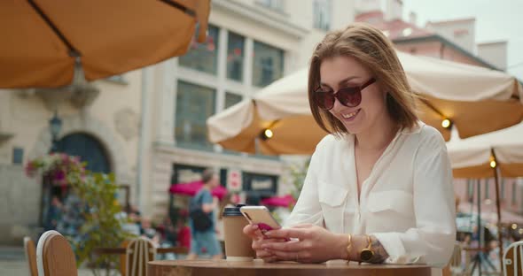 The Smiling Girl is Sitting at a Table in a Cafe and Texting on the Phone