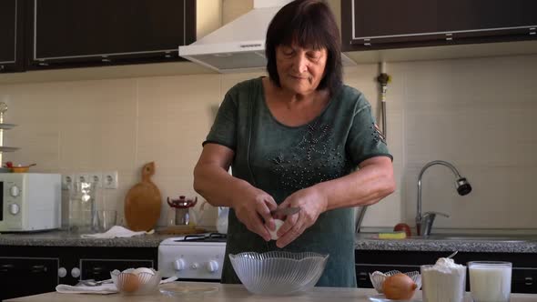 Happy 60s Old Lady Breaking Eggs Into Bowl at Kitchen Table Happy Smiling