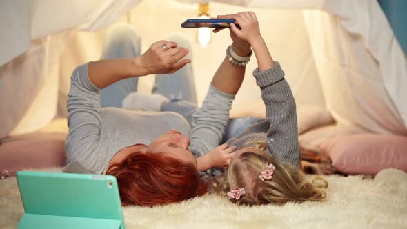 Relaxed Mother and Little Daughter Taking Selfie on Smartphone Lying in Cozy Tent in Living Room