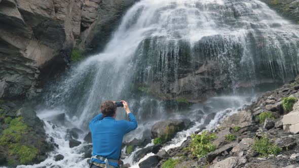 Man Holds Phone and Makes Video Standing Near Waterfall